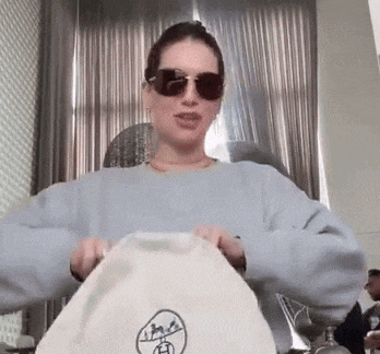 a woman wearing sunglasses and a light blue sweater is holding a white bag .