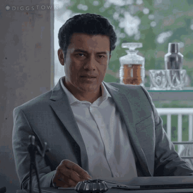 a man in a suit and white shirt is sitting at a desk with a laptop and a bottle of whiskey behind him