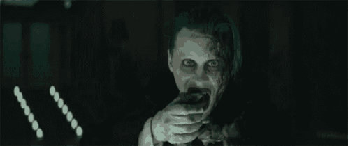 the joker from suicide squad is pointing at the camera with a knife in his mouth .