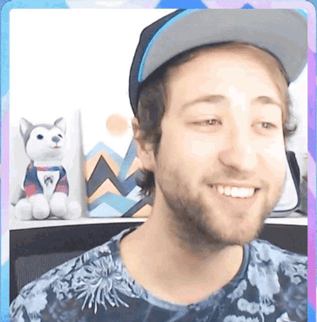 a man wearing a hat and a floral shirt smiles in front of a stuffed dog