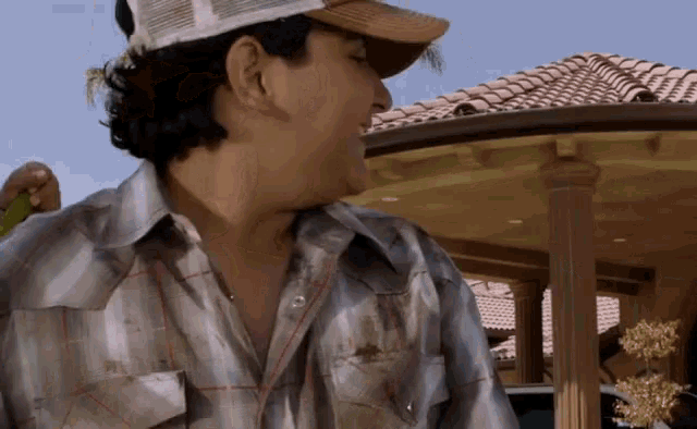 a man wearing a plaid shirt and a hat stands in front of a house