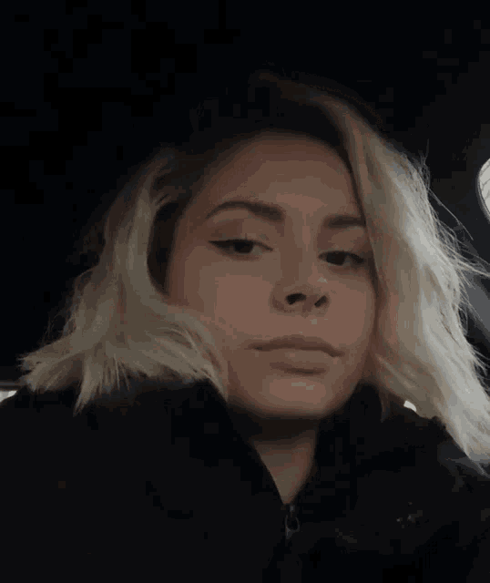 a woman with blonde hair wearing a black jacket looks at the camera
