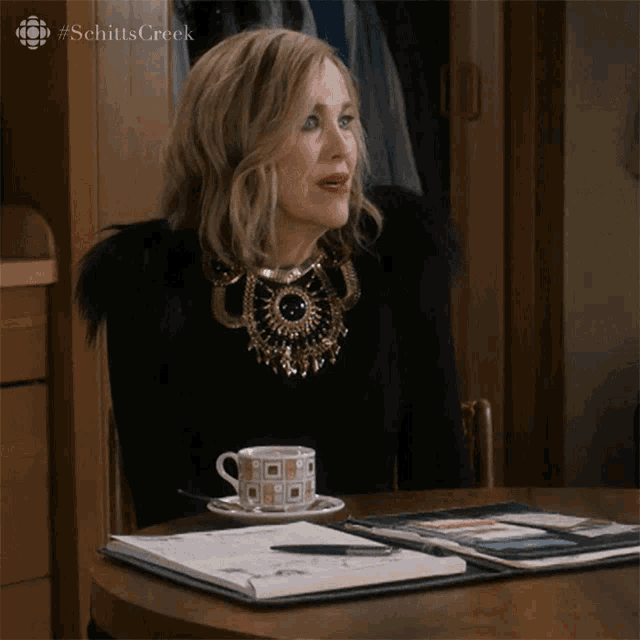 a woman is sitting at a table with a cup of coffee and a notebook with the word schitts creek on it