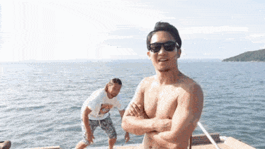 a shirtless man wearing sunglasses stands on a dock while another man stands in the water