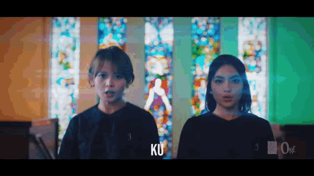 two women are standing in front of a stained glass window and the letters ku are visible