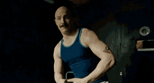 a man with a mustache and a blue tank top is flexing his muscles in a dark room .