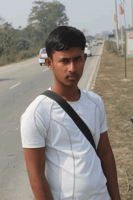 a young man in a white shirt with a black strap around his shoulder stands in front of a highway