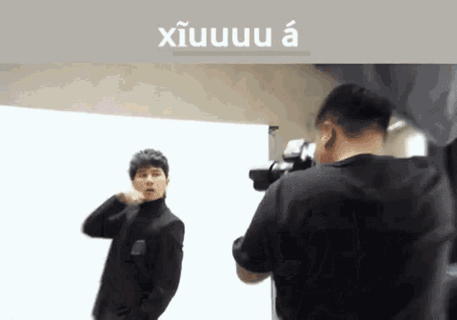 a man is taking a picture of another man in front of a white wall with a camera .