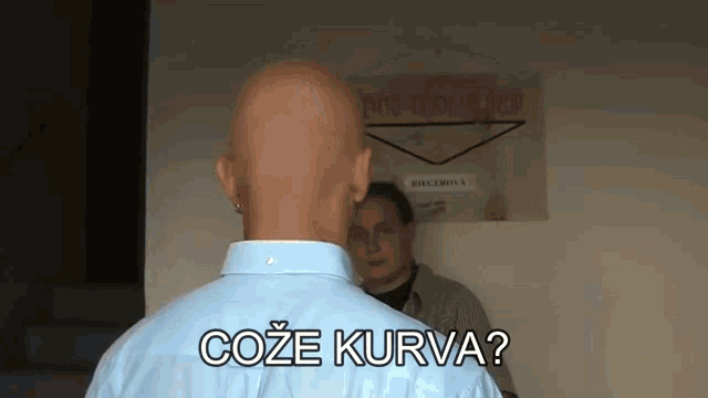 a bald man in a blue shirt is standing in front of a sign that says coze kurva