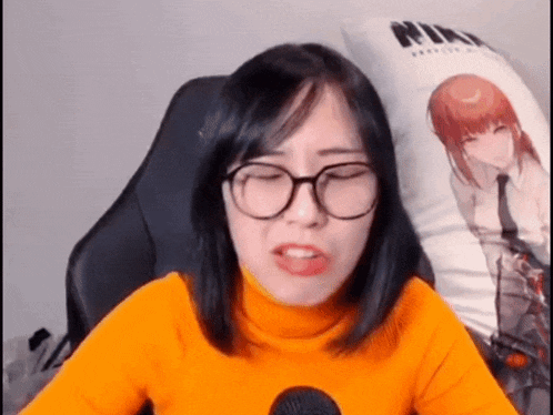 a woman wearing glasses and an orange turtleneck is sitting in a chair with a pillow .