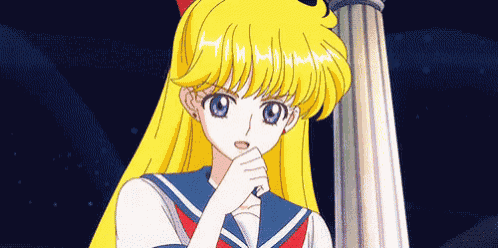 a girl with long blonde hair and blue eyes is standing next to a pillar and holding her hand to her mouth .