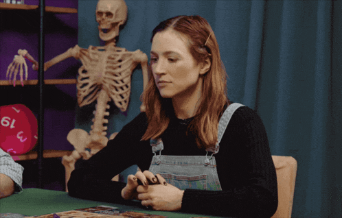 a woman sits at a table in front of a skeleton and a dice that says 19