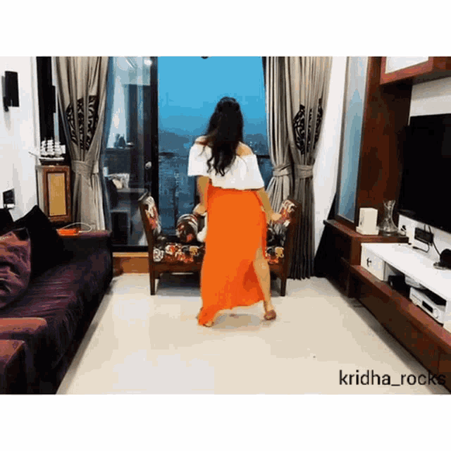 a woman in an orange skirt is dancing in a living room with the name krishna_rocks at the bottom
