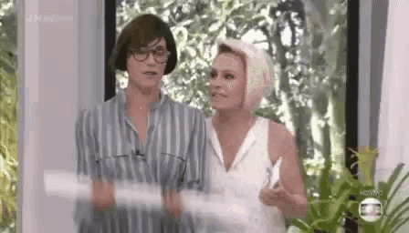 two women are standing next to each other in front of a window and talking .