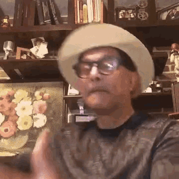 a man wearing a hat and glasses is making a funny face in front of a bookshelf .