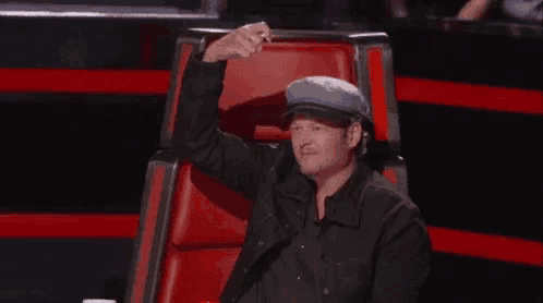 a man wearing a hat is sitting in a red chair and raising his hand .