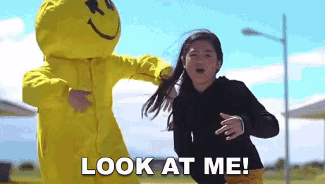 a girl in a black hoodie is standing next to a yellow smiley face mascot and says `` look at me '' .