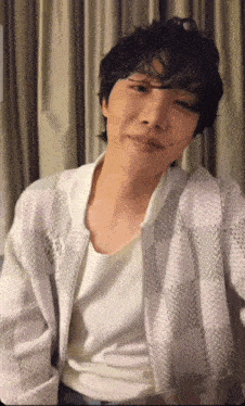 a man with curly hair is sitting in front of a window wearing a white shirt and a white jacket .