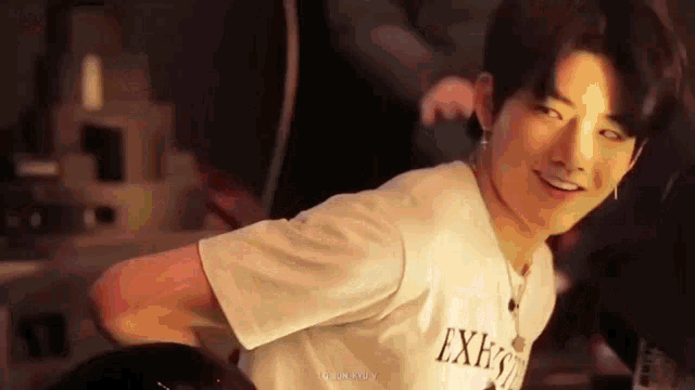 a young man in a white t-shirt is smiling and looking at the camera while standing in front of a mirror .