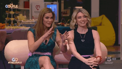 two women sit on a couch in front of a sign that says the couple on it