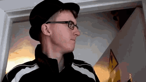 a man wearing a hat and glasses is standing in front of a yellow sign that says ' warning ' on it
