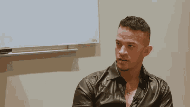 a man in a leather shirt is sitting in front of a whiteboard