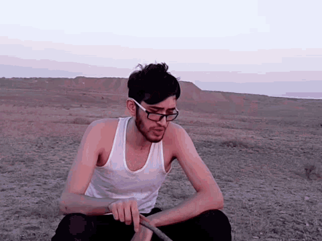 a young man wearing glasses and a white tank top is sitting in the desert