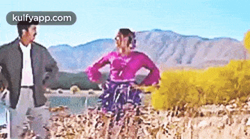 a man and a woman are standing in a field with mountains in the background . the woman is wearing a pink dress .