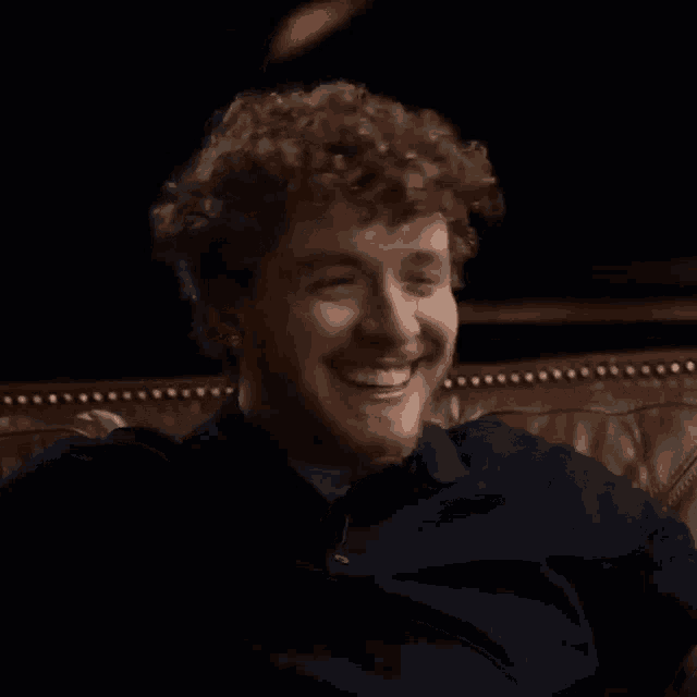 a man with curly hair and a beard is sitting on a couch and smiling .