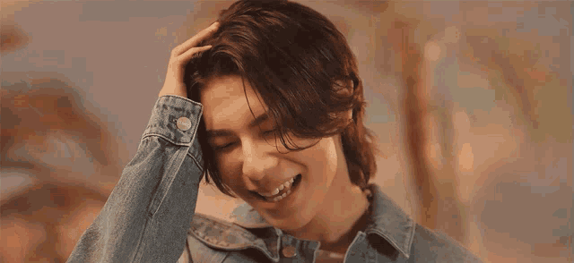 a young man wearing a denim jacket is smiling with his hand on his head