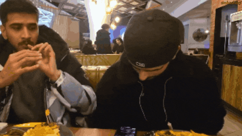 two men are sitting at a table eating food and one of them is wearing a hat that says cag
