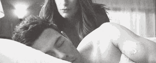 a black and white photo of a woman standing next to a man sleeping on a bed .