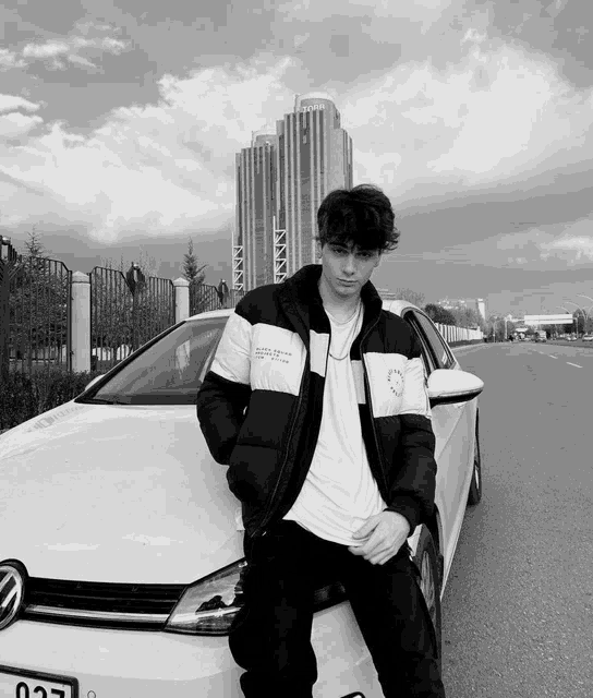 a black and white photo of a young man standing next to a white car with a license plate that says 027