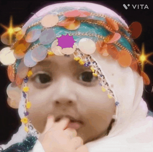 a baby wearing a hijab and a headband with sequins on it is licking her fingers .