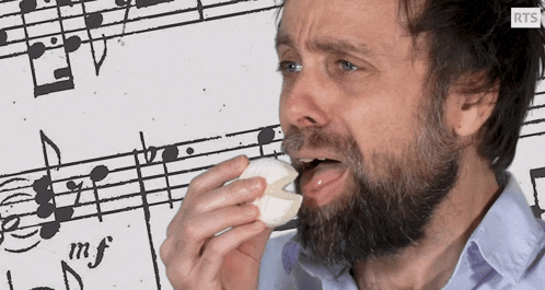 a man with a beard is eating a piece of food in front of a sheet of music with the letters rts on it