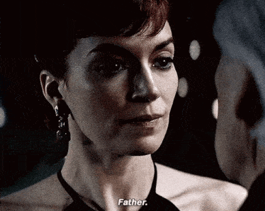 a woman in a black dress is talking to a man in a dark room and says father .