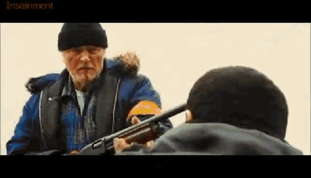 a man with a beard is holding a shotgun in front of another man