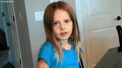 a little girl in a blue shirt is making a funny face while sitting at a table .