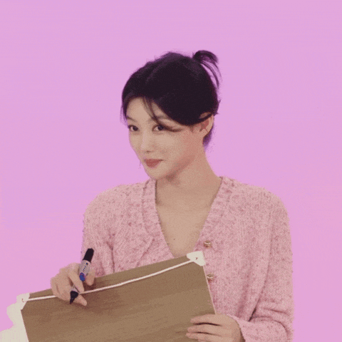 a woman in a pink sweater is writing on a piece of paper with a marker