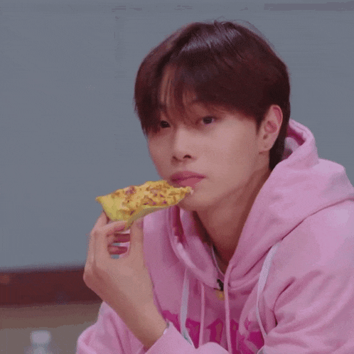 a young man in a pink hoodie is eating a piece of pizza