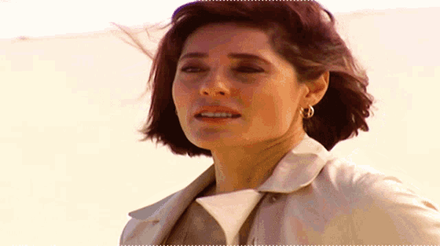 a woman wearing a tan trench coat and hoop earrings looks at the camera