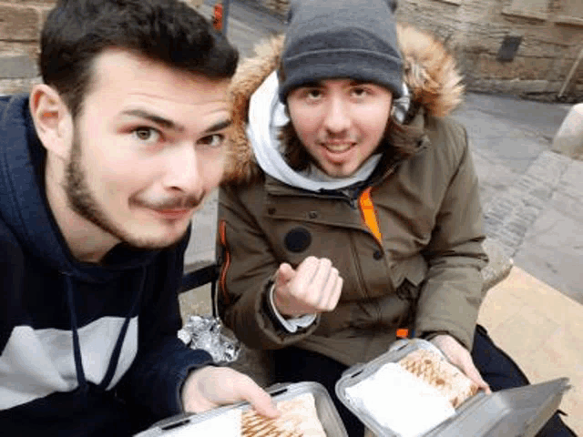 two men are sitting next to each other on a bench eating food from plastic containers .
