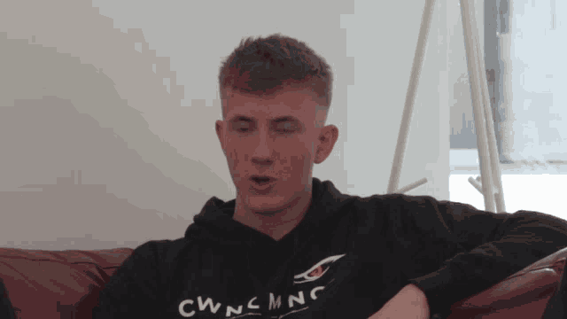 a young man sitting on a couch wearing a black sweatshirt that says cwncmnc