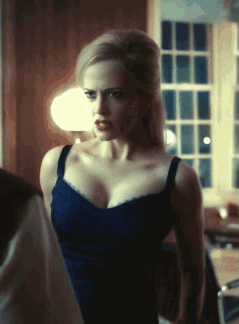 a woman in a blue dress with a very large breast is standing in front of a window
