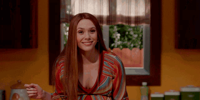 a woman with long red hair is sitting at a table in front of a window smiling .