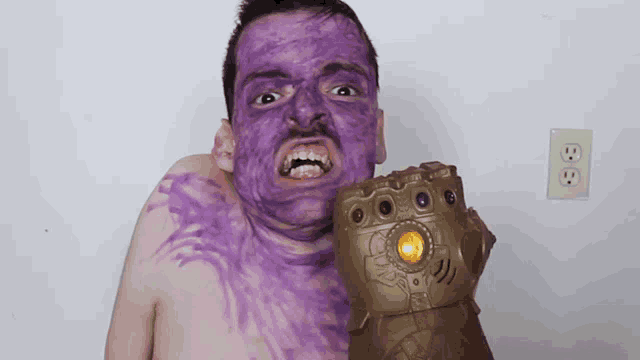 a man with purple paint on his face is holding a glove that says infinity gauntlet