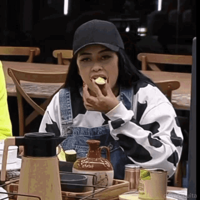 a woman wearing overalls and a black hat is eating a piece of food