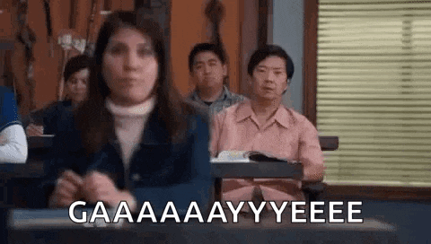 a woman is sitting at a desk in a classroom with other people and making a funny face .