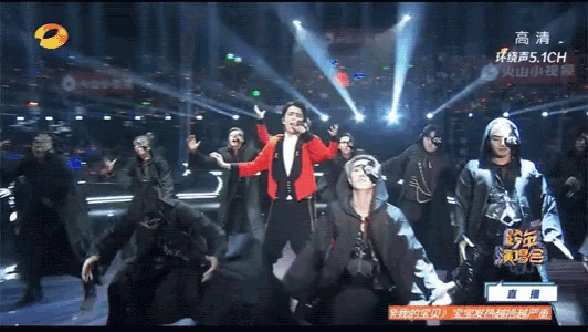 a man in a red jacket is singing into a microphone on a stage surrounded by dancers