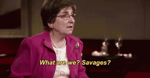 an elderly woman wearing glasses and a pink jacket is sitting at a table and asking what are we ? savages .